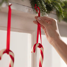 A hand hangs a red and white candy cane on a red ribbon on a white surface, with Command™ Mini Hooks, transparent, with transparent strips, 17006CLRFGN by 3M Deutschland GmbH. Another candy cane is already hanging next to it on green pine garland.