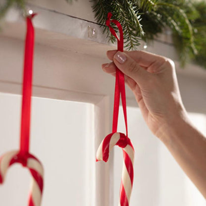 A hand hangs a red and white candy cane on a red ribbon on a white surface, with Command™ Mini Hooks, transparent, with transparent strips, 17006CLRFGN by 3M Deutschland GmbH. Another candy cane is already hanging next to it on green pine garland.