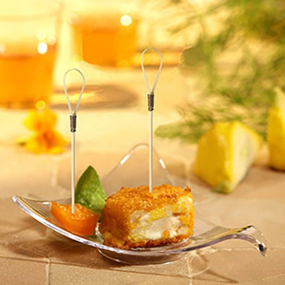 A glass bowl presents a breaded, fried appetizer cube with a PAPSTAR finger food picker "Loop" (10 cm), an orange segment, and a green garnish. In the background, lemon wedges and two drinks from PAPSTAR GmbH blur.