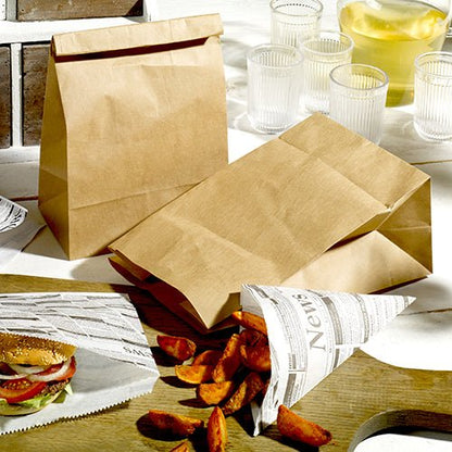 PAPSTAR 250 block bottom bags made of brown kraft paper from PAPSTAR GmbH stand with newspaper-wrapped food on wood, along with glasses, a jug with a light drink, as well as fries and a burger in the background.