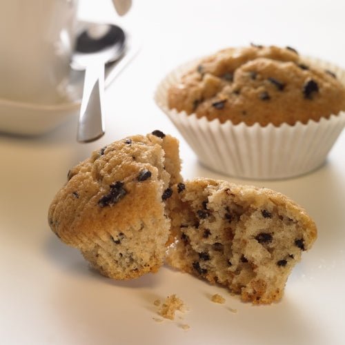 In the foreground, a halved chocolate chip muffin is visible, with crumbs scattered over it. In the background, there is another muffin in a PAPSTAR 1000 baking cup round of PAPSTAR GmbH, next to a white cup with saucer and spoon on a white surface.