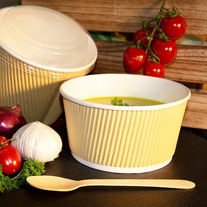A PAPSTAR round soup cup, 11 cm, brown/white, Ripple Wall (25 pieces) from PAPSTAR GmbH is filled with yellow soup and herbs, next to it a wooden spoon, cherry tomatoes on the vine, garlic, an onion, and parsley on a black surface.