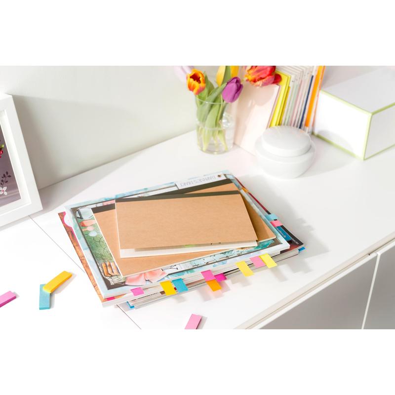 A stack of books and a brown notebook with colorful 3M Post-it® Page Markers from the Poptimistic Collection lie on a white desk. In the background, there are tulips in a vase, more books, and a white round object.