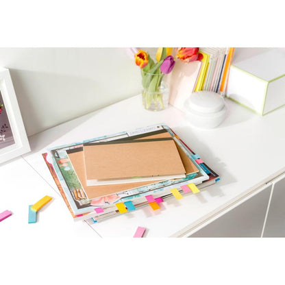 A stack of books and a brown notebook with colorful 3M Post-it® Page Markers from the Poptimistic Collection lie on a white desk. In the background, there are tulips in a vase, more books, and a white round object.
