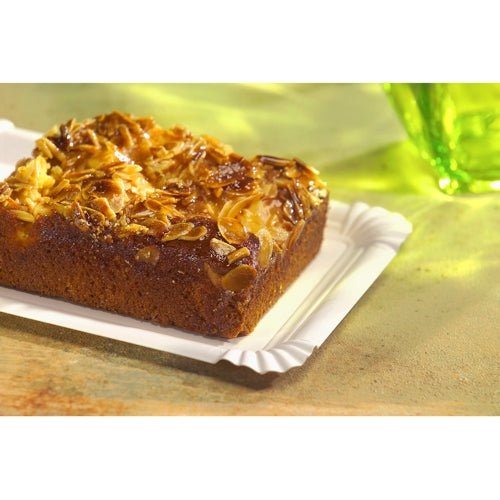 A rectangular almond cake rests on a PAPSTAR GmbH PAPSTAR 250 plate, cardboard "pure" square white with decorative edges. In the background, a green glass and a textured surface in warm tones can be seen.