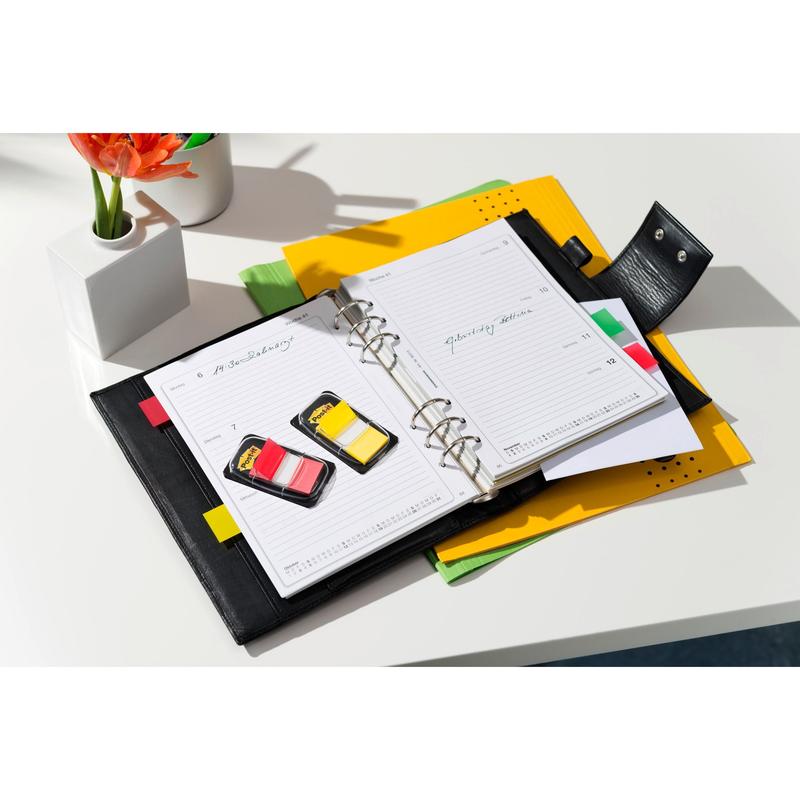 An open black planner with lined pages lies on a white desk, surrounded by colorful folders, 3M Deutschland GmbH Post-it® Index Tabs, Standard (25.4 mm x 43.2 mm), and a small white vase with an orange flower.