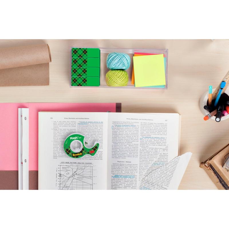 A desk with an open book, Scotch® Magic™ Invisible Tape Value Pack (24 rolls, 3M Deutschland GmbH), colored paper, sticky notes, string, eraser, a cup with pens and pencils, as well as stacked books and rolled brown paper.