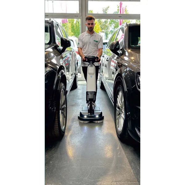 A person in a white shirt uses the Arcora Tiger 40 floor cleaning machine from Arcora International GmbH between two black cars in a bright interior with large windows, ensuring a professional cleaning.