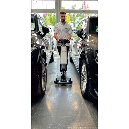 A person in a white shirt uses the Arcora Tiger 40 floor cleaning machine from Arcora International GmbH between two black cars in a bright interior with large windows, ensuring a professional cleaning.