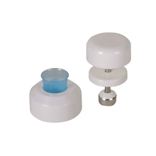 Against a white background, two white plastic kitchen tools can be seen. The tool on the left has a cylindrical shape with a blue plastic insert, while the tool on the right is an AMPri tablet crusher for medicine cups by AMPri Handelsgesellschaft mbH, which consists of two white discs connected by a stainless steel rod with a metal base.