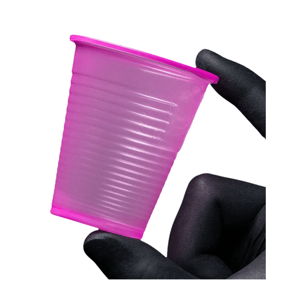 A small, pink AMPri Dental PP mouth rinse cup made of polypropylene is held between two gloved fingers against a white background. This disposable mouth rinse cup from AMPri Handelsgesellschaft mbH, often found in dental practices, features a textured surface with horizontal grooves.