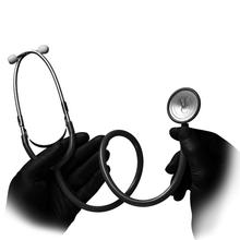 A pair of black-gloved hands holds the AMPri MED-COMFORT PVC flathead stethoscope from AMPri Handelsgesellschaft mbH against a white background. The partially coiled stethoscope is made of polyvinyl chloride. One hand holds the ear tips and the other the diaphragm.