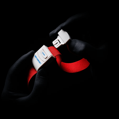 Gloved hands, reminiscent of an AMPri MED-COMFORT tourniquet, grip the buckle of a bright red safety belt against a shaded background. The word "Blue" and the letter "B" can be seen on the buckle.