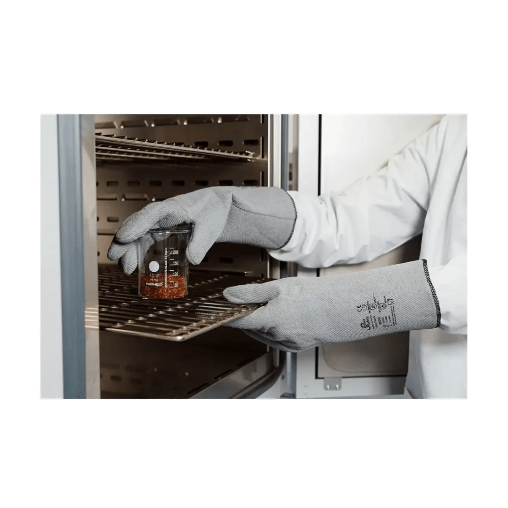 A person wears AMPri SolidSafety heat protection gloves L - XXL from AMPri Handelsgesellschaft mbH and a lab coat and carefully places a glass beaker with reddish liquid on a metal stand in an industrial oven. The focus is on the hands and the beaker, emphasizing the maximum heat protection, with the shelves inside the device visible.
