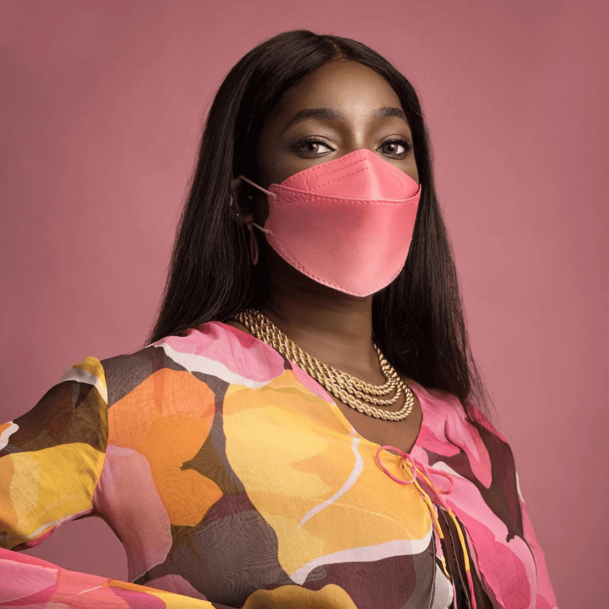 A person wears a particle-filtering Altruan designer face mask (FFP2 standard) in a stylish pink hue and a colorful top with an abstract pattern in front of a pink background. She has long, dark hair and wears multiple gold necklaces.