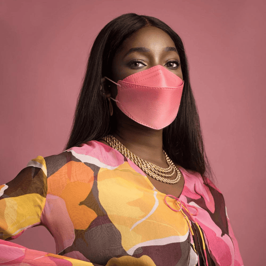 A person wears a particle-filtering Altruan designer face mask (FFP2 standard) in a stylish pink hue and a colorful top with an abstract pattern in front of a pink background. She has long, dark hair and wears multiple gold necklaces.