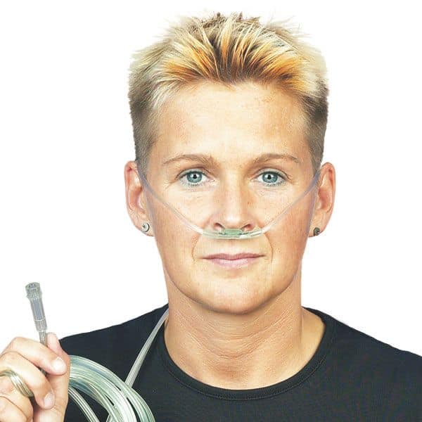 A person with short hair is using the DCT oxygen nasal cannula Soft for adults from Servoprax GmbH for oxygen support. They are holding the O2 connecting tube and wearing a black shirt in front of a plain white background.