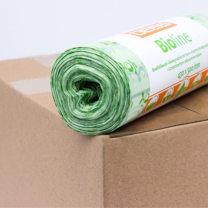 A roll of green compostable waste bags is placed on a cardboard box. The label on the roll is partially visible and bears the print "EMIL DEISS KG (GmbH + Co.)". These 100% compostable bags have a floral design and measure 430 x 500 mm.