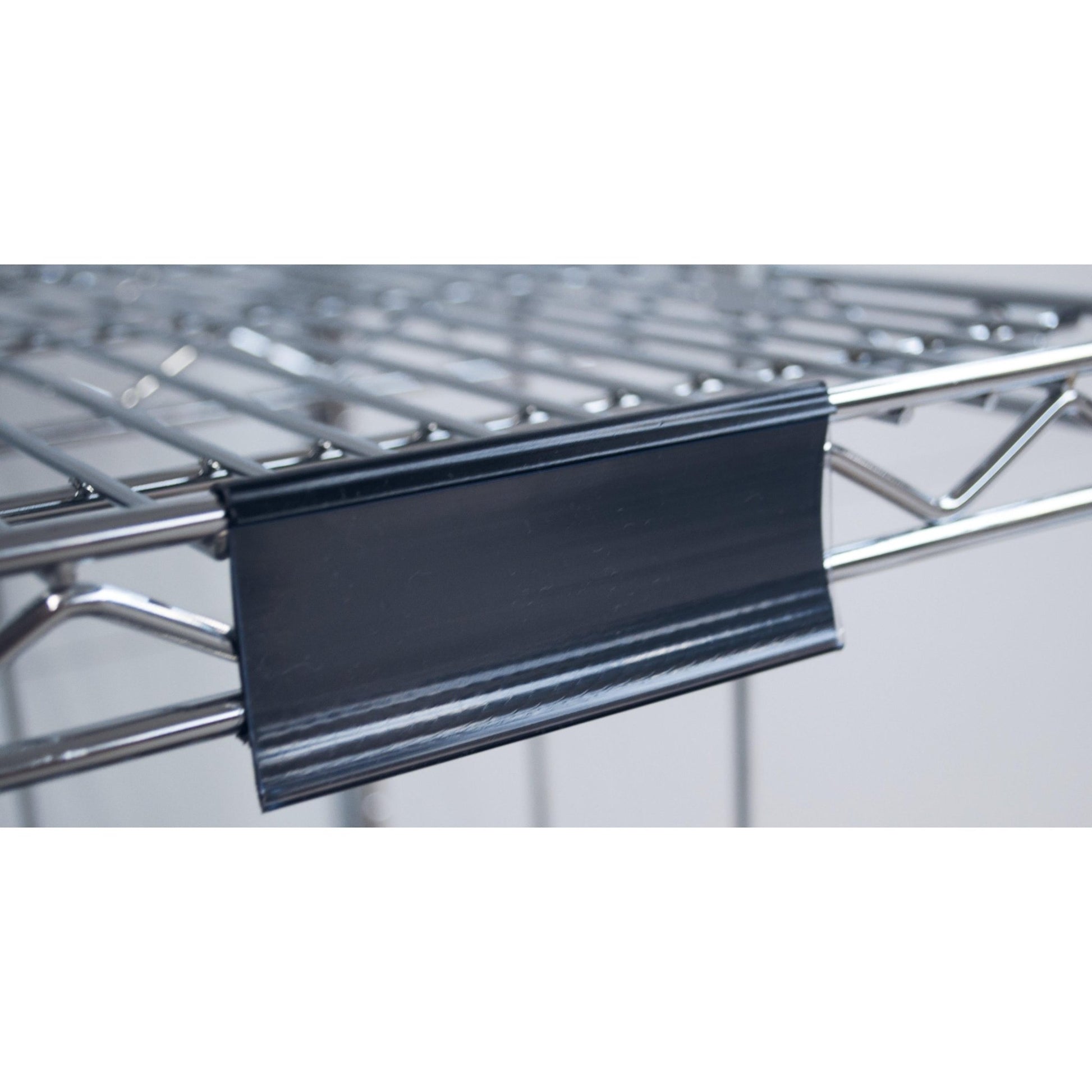 Close-up of a Novocal label frame with paper tags (approx. 100 x 44 mm, 1 piece) attached to a metal wire shelf, with the black plastic label holder on the front edge; the background is blurred.