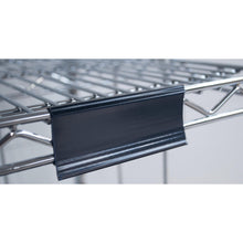Close-up of a Novocal label frame with paper tags (approx. 100 x 44 mm, 1 piece) attached to a metal wire shelf, with the black plastic label holder on the front edge; the background is blurred.