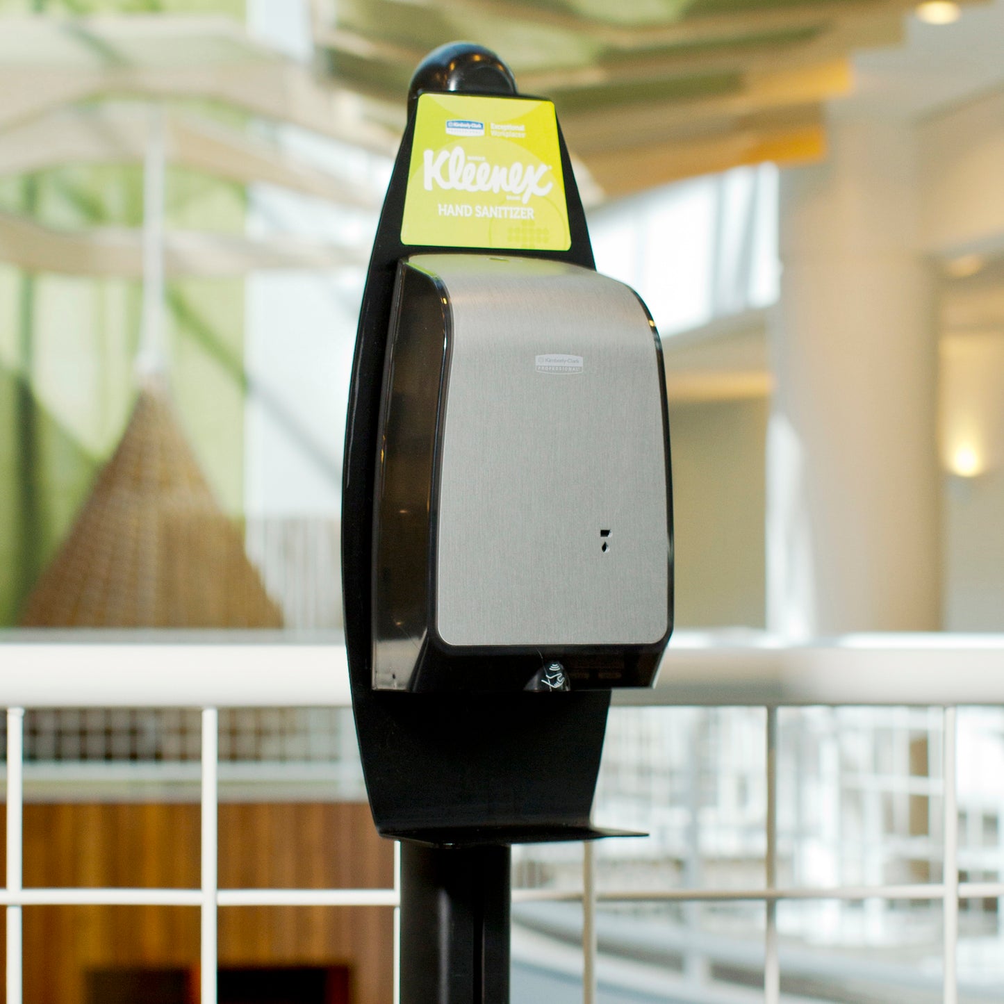 A Kimberly-Clark GmbH Electronic Lotion Stand (Black, 1 dispenser) is placed indoors near a white metal railing for hygienic hand cleaning, with blurred architectural details in the background.