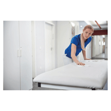 A woman in a blue shirt and white pants cleans a white examination table with the Bacillola® Zero Tissues Quick-Disinfection Wipes (1 piece) by Paul Hartmann AG for eco-friendly disinfection in a bright medical environment.
