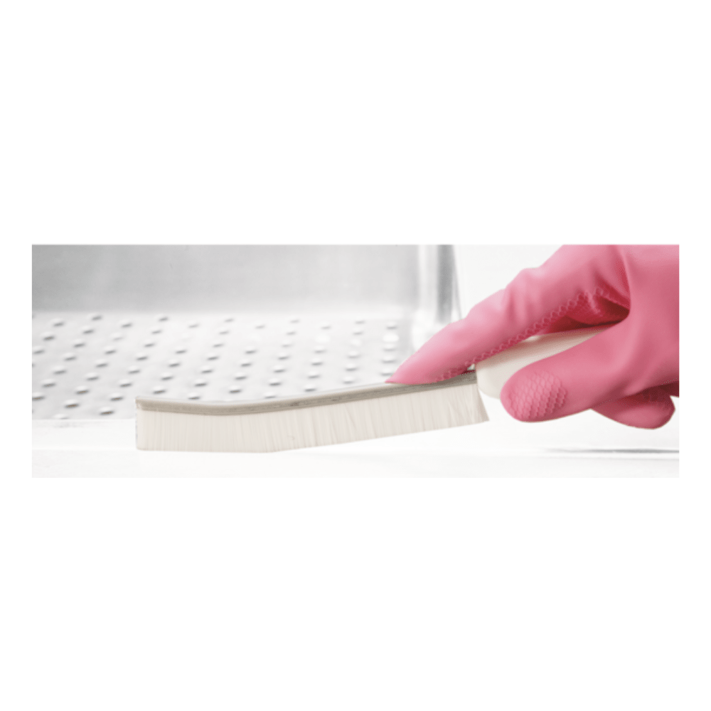A hand with a pink rubber glove holds a Haug Bürsten KG narrow joint brush PP ø 0.40 mm Hard with white bristles. In the background, a partially visible metal sink can be seen, highlighting its purpose for cleaning narrow areas.