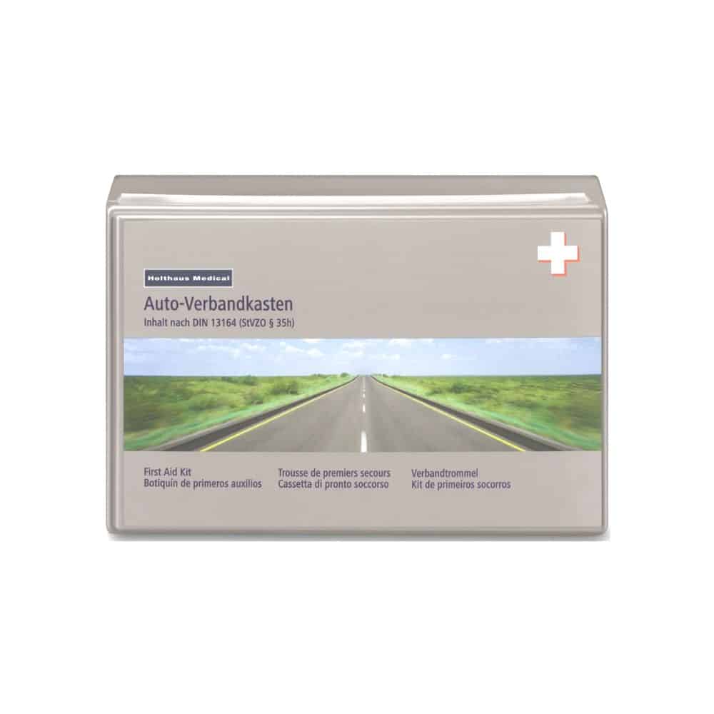 A rectangular plastic first-aid kit with a transparent cover from Holthaus Medical GmbH & Co. KG. The label is multilingual and features a road stretching to the horizon with a white cross in the upper right corner. The text “Holthaus Medical Classic Car First Aid Kit DIN 13164” indicates its durable, impact-resistant design.