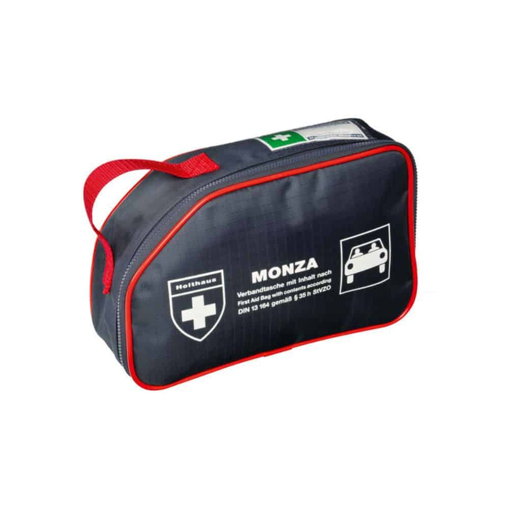 Displayed is a compact, black first aid kit with red piping and a red carrying strap. The kit bears the inscription “Holthaus Medical MONZA Verbandtasche Auto” from Holthaus Medical GmbH & Co. KG. On the front, there is a white cross symbol and a car symbol, as well as some German text details, identifying it as an ideal car first aid kit for on the go.