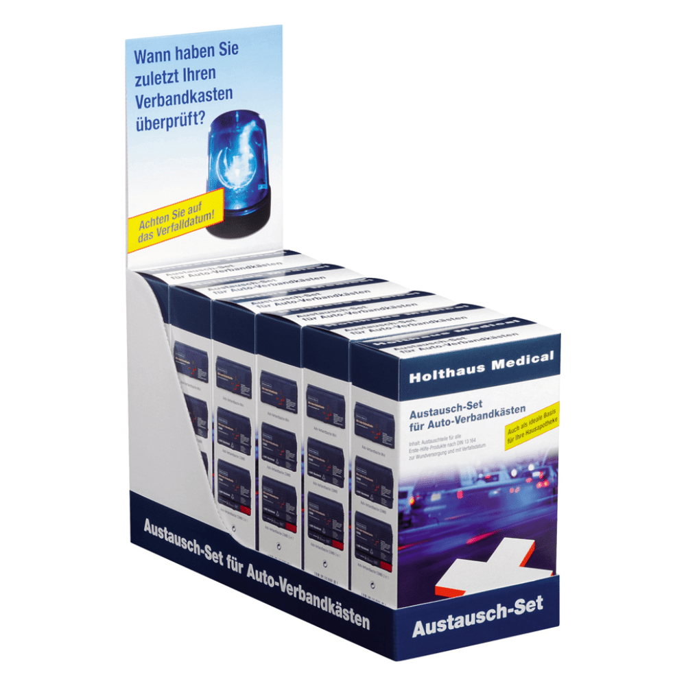 An exhibition stand presents several boxes of the Holthaus Medical replacement set for car first aid kits | Package (1 piece) by Holthaus Medical GmbH & Co. KG. The top sign urges customers with a blue siren image and German text to check their first aid equipment. The stand features a white base with blue accents and additional German text.