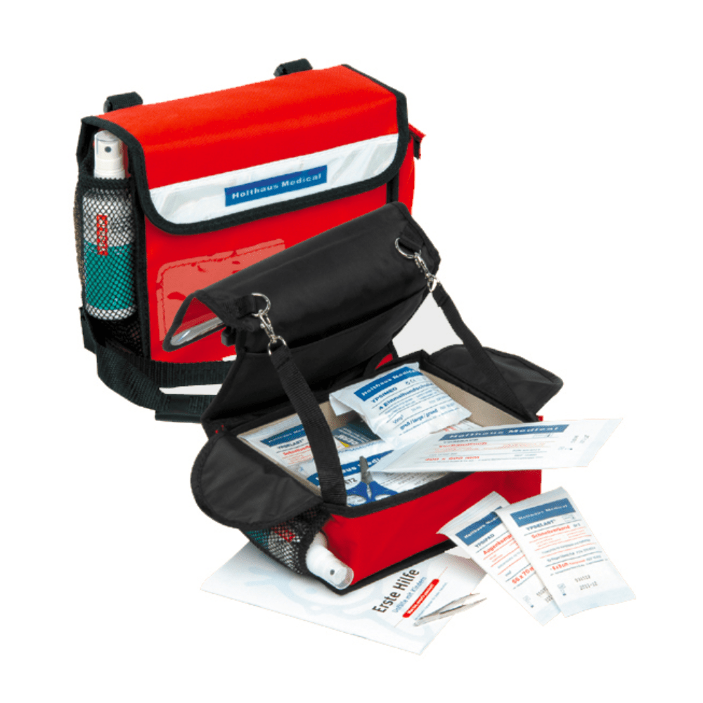 A red Holthaus Medical first aid kit for kindergarten by Holthaus Medical GmbH & Co. KG is open and displays a selection of first aid items, including bandages, dressings, a first aid booklet, and a small bottle. The bag has a black flap and side pockets made of mesh fabric. It is ideally suited as a first aid kit in environments such as kindergartens.