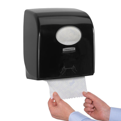 A person pulls paper towels from a Kimberly-Clark GmbH Aquarius™ Slimroll™ roll towel dispenser - Black, which is wall-mounted. Only the hands are visible, demonstrating the hygienic paper towel dispensing through the oval window.