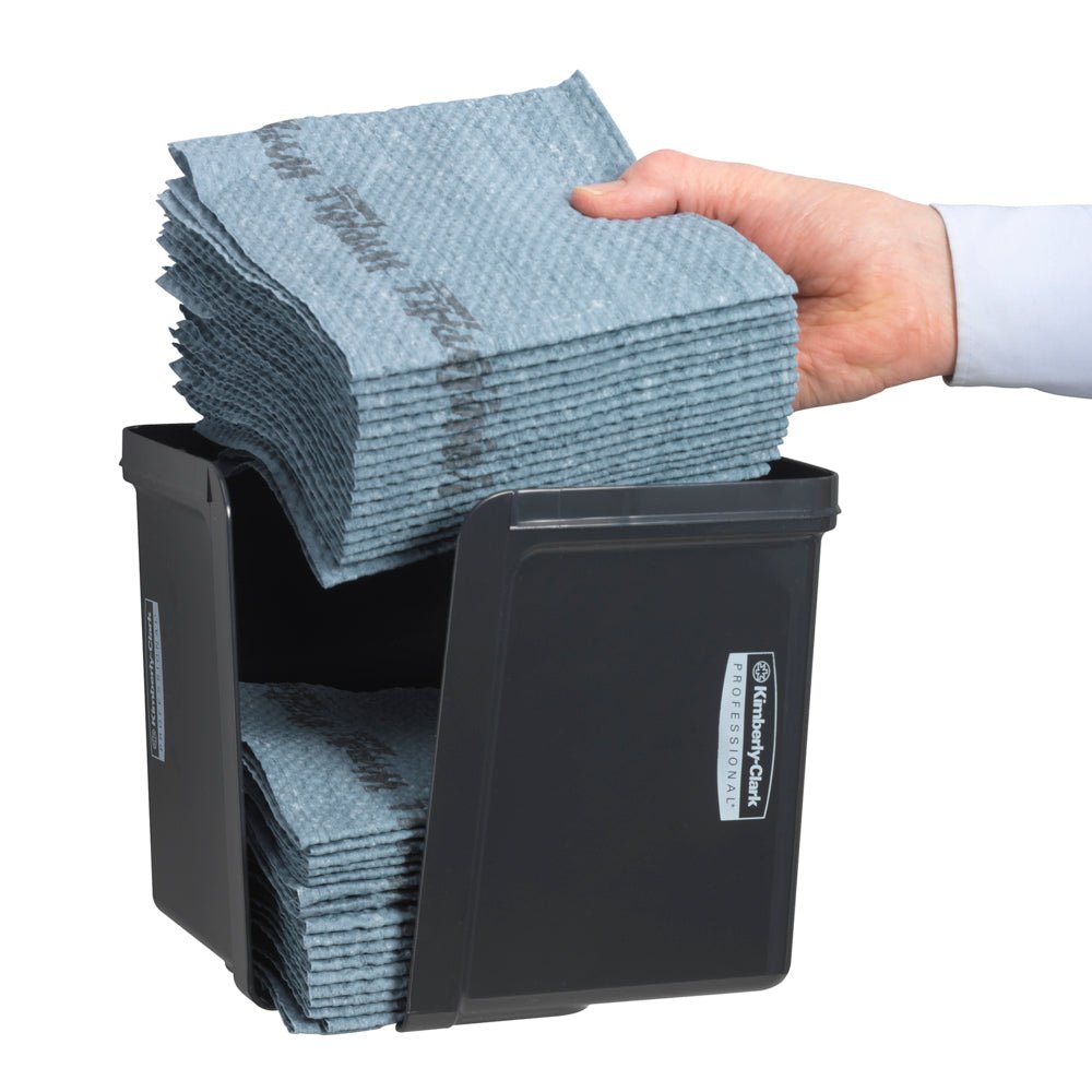 A hand inserts quarter-folded wipes into the gray Kimberly-Clark Professional™ wipe dispenser from Kimberly-Clark GmbH - for hygienic cloth removal. The neatly folded wipes and the front label are visible.