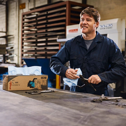 A man in a dark overall wipes a tool with a blue cloth on a workshop table with WypAll L40 wipes, industrial equipment, and Hostess™ NATURA Toilet Paper (Single Sheet System, White, 32 x 500 sheets) from Kimberly-Clark GmbH.