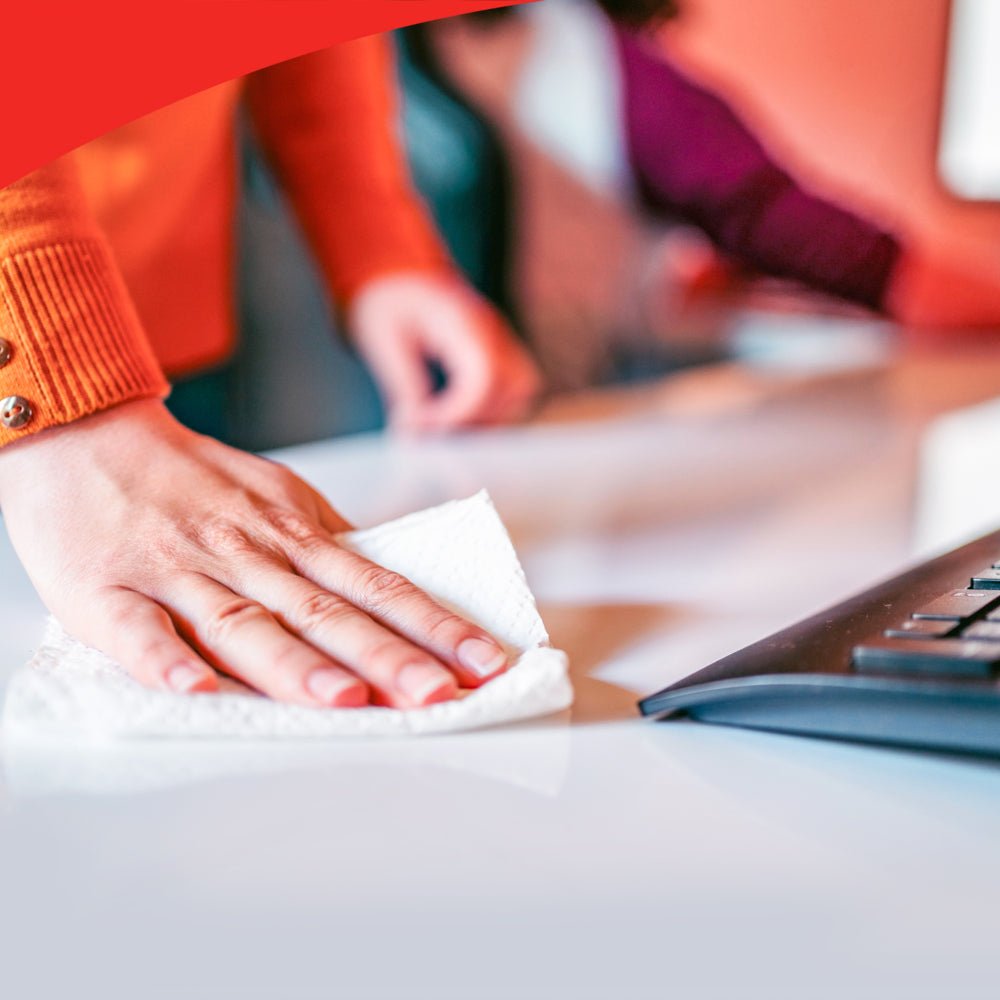 A person in an orange sweater wipes a white desk with a cloth, likely from the WypAll® X60 General Clean pop-up box from Kimberly-Clark GmbH, next to a keyboard. Another person is blurred in the background, and a red graphic is visible in the upper left corner.