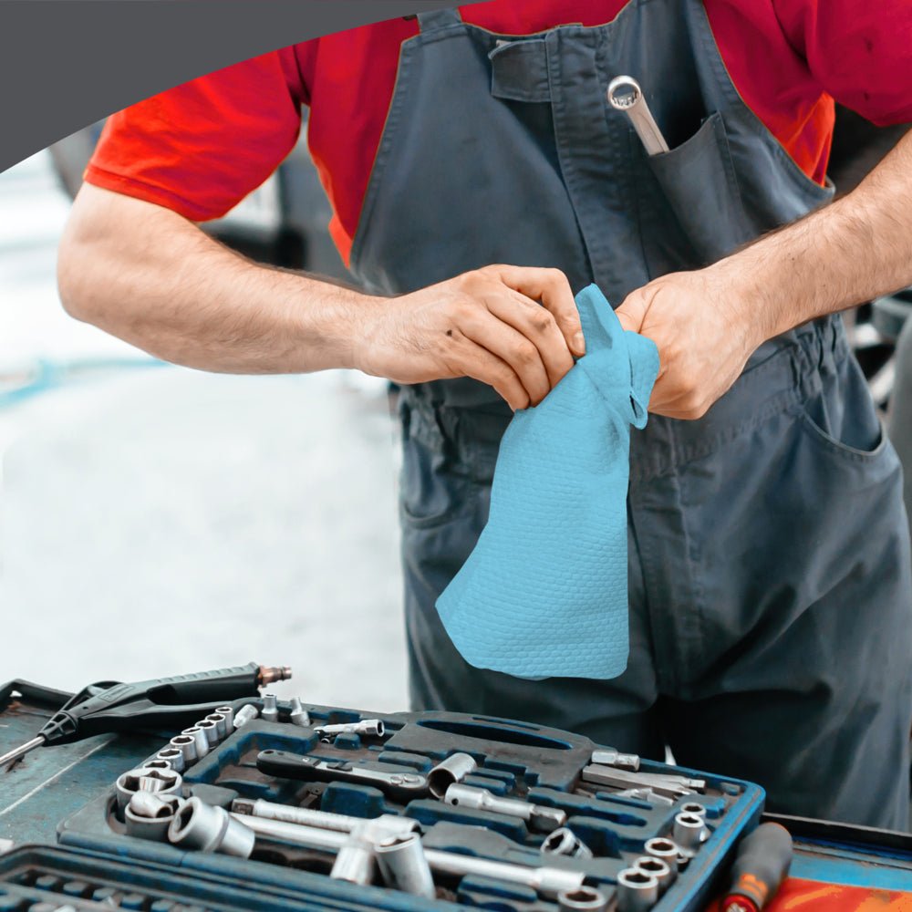 A person in a gray overall and red shirt wipes their hands with a WypAll® X70 PowerClean - Grab Box (Blue) from Kimberly-Clark GmbH next to a tool-laden workbench, ensuring professional workplace cleaning.
