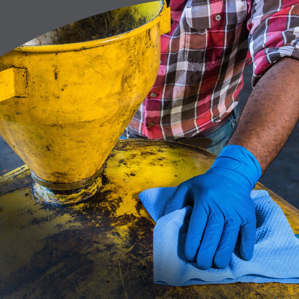 In a red checkered shirt and blue gloves, someone wipes a yellow industrial container with a WypAll® X80 PowerClean cloth from the Steel Blue pull-up box by Kimberly-Clark GmbH, using one of the 5 boxes with 80 cloths per carton.