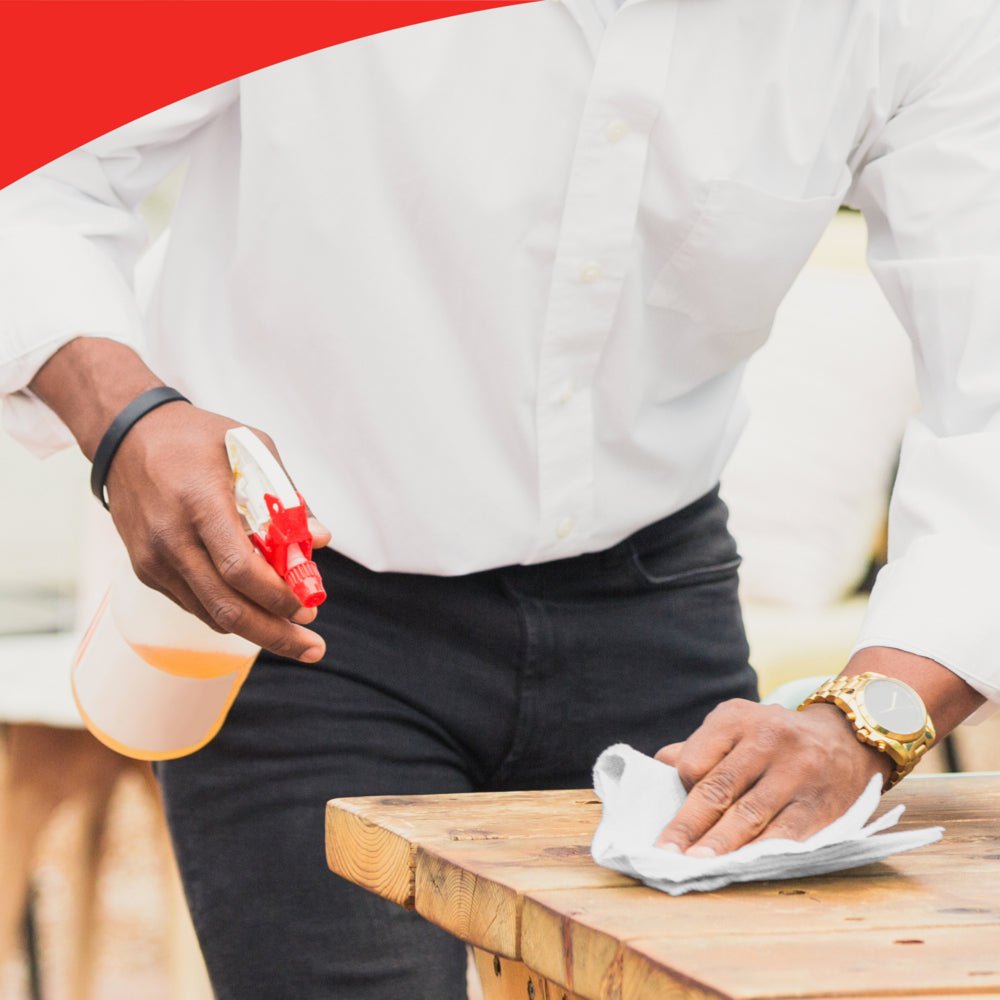 A person in a white shirt and black pants uses a WypAll® X60 General Clean large roll from Kimberly-Clark GmbH and a spray bottle to thoroughly clean a wooden table.