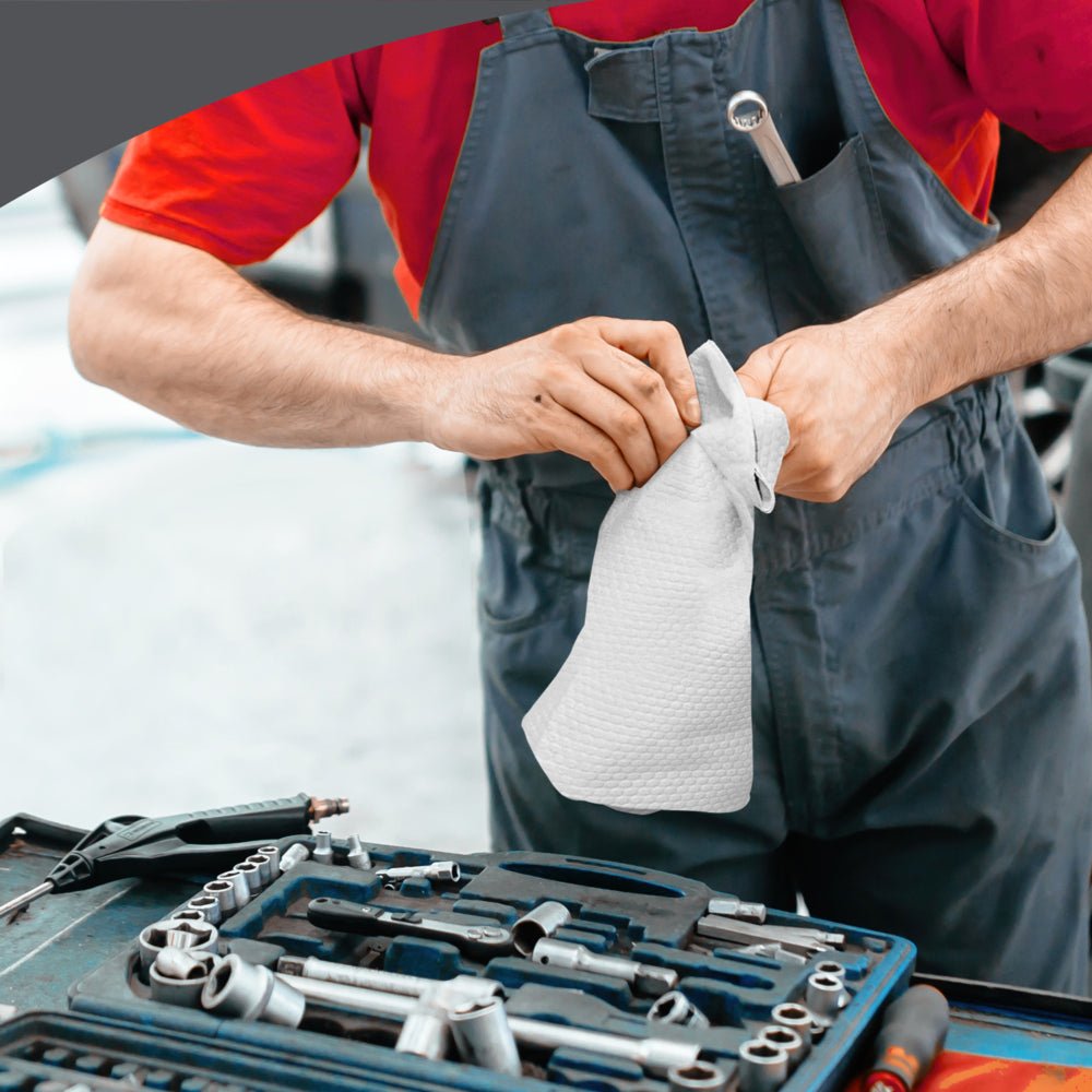 A person in a red shirt and gray overalls cleans their hands with a WypAll® X80 PowerClean Quarter-Folded white cloth from Kimberly-Clark GmbH over a workbench, next to an open toolbox with various tools and wrenches.