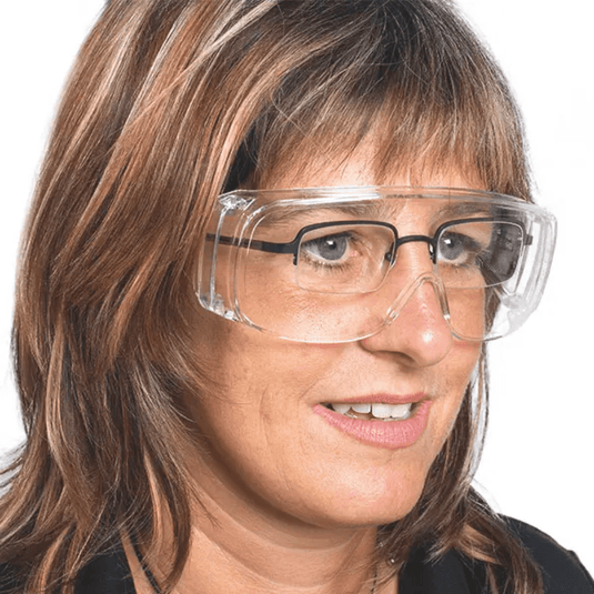 A person with shoulder-length hair is wearing a transparent Mediware protective eyewear made of polycarbonate from Servoprax GmbH over regular glasses. They are looking slightly to the side against a plain white background.