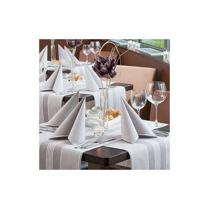 The dining table is elegantly set with folded napkins, wine glasses, water glasses, and decorative flowers. A bright tablecloth from the ROYAL Collection of PAPSTAR GmbH complements the matching napkins. The noble ambiance, complemented by a tasteful PAPSTAR table runner fabric-like PV-Tissue Mix “ROYAL Collection” measuring 24 m x 40 cm, is perfect for a refined meal.