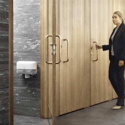 A woman in black business attire opens a wooden door in a restroom, revealing walls clad in gray marble tiles. A white hand dryer and a Tork 558040 double roll dispenser for coreless Midi toilet paper Elevation T7 from TORK – Essity Professional Hygiene Germany GmbH are mounted on a wall, highlighting the eco-friendly design.