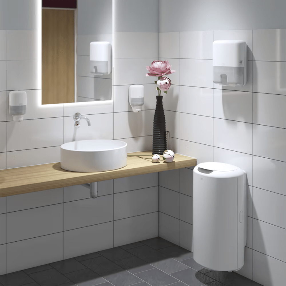 A modern bathroom presents a round white sink on a wooden countertop, adorned with a tall vase of pink flowers. The Tork 561000 Mini Dispenser for Liquid Soap Elevation S2 from TORK - Essity Professional Hygiene Germany GmbH provides function and style under the large illuminated mirror. Soap dispensers on the white tiled walls emphasize the elegance design, while a sleek white trash bin stands on the contrasting dark floor tiles.