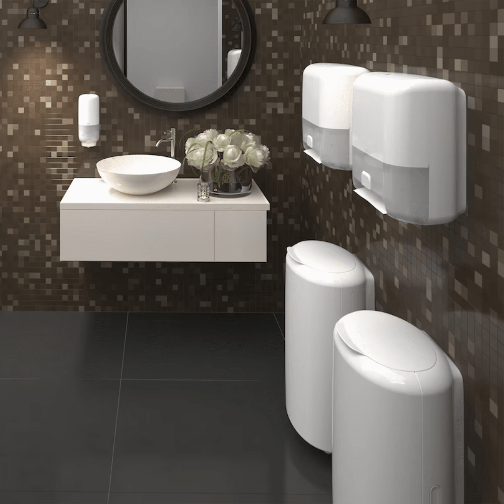 A modern bathroom with dark mosaic tiles on the walls and a black tiled floor features a round mirror above a wall-mounted sink with a white countertop basin. Two Tork 561600 Skincare Dispensers with Intuition Sensor by TORK – Essity Professional Hygiene Germany GmbH ensure touchless dispensing, and two waste bins are placed on the right. A vase with white flowers adorns the sink.