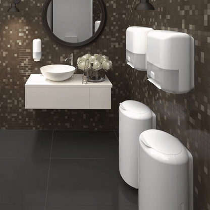A modern bathroom impresses with an upscale design featuring a dark-tiled wall and a floating white sink under a round mirror. On the right, the Tork 563000 Waste Bin Elevation 50 Liter B1 by TORK – Essity Professional Hygiene Germany GmbH offers elegant waste solutions, while a bouquet of white flowers emphasizes the elegance of the countertop.