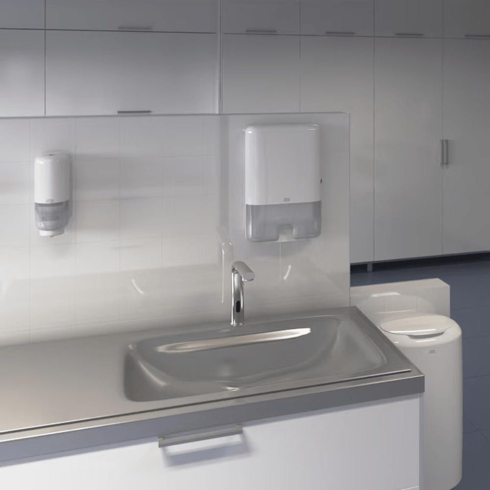 A modern sink with a stainless steel plate and faucet is a highlight of the Elevation design. On the tiled wall above the sink, a soap dispenser and a paper towel dispenser are mounted, while in the background, a Tork 563000 Waste Bin Elevation 50 Liter B1 by TORK – Essity Professional Hygiene Germany GmbH can be seen.