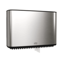 A Tork Xpress dispenser for mini-jumbo toilet paper, part of the Image Line, is elegantly mounted on the wall. This stainless steel dispenser has a sleek rectangular design, with a small section of white paper visible at the bottom. The brand "TORK – Essity Professional Hygiene Germany GmbH" is clearly visible on the front.