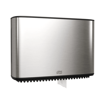 A Tork Xpress dispenser for mini-jumbo toilet paper, part of the Image Line, is elegantly mounted on the wall. This stainless steel dispenser has a sleek rectangular design, with a small section of white paper visible at the bottom. The brand "TORK – Essity Professional Hygiene Germany GmbH" is clearly visible on the front.