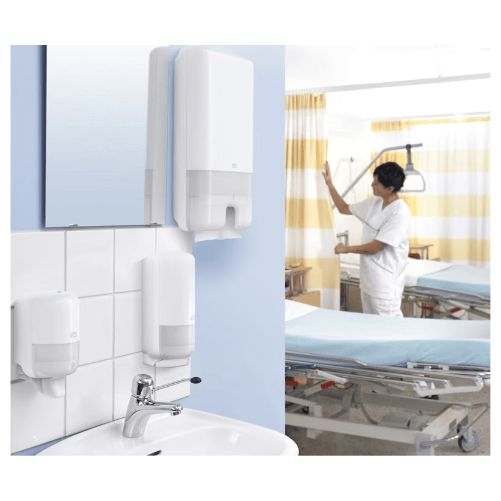 In a hospital room, a caregiver adjusts the curtain next to a patient's bed. In the foreground, Tork Xpress® 471093 Multifold Towels Universal H2 1-ply from TORK – Essity Professional Hygiene Germany GmbH are placed next to universal soap. The bed is covered with blue sheets and surrounded by important medical equipment.