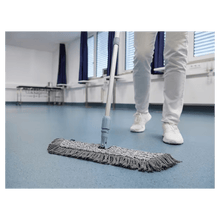 A person in white shoes and pants is mopping a blue floor with a Vileda GmbH Professional Swep Duo r-MicroCombi Mop - 50 cm in a room with closed blinds and blue curtains. A folding table stands nearby and a radiator is under the windows.